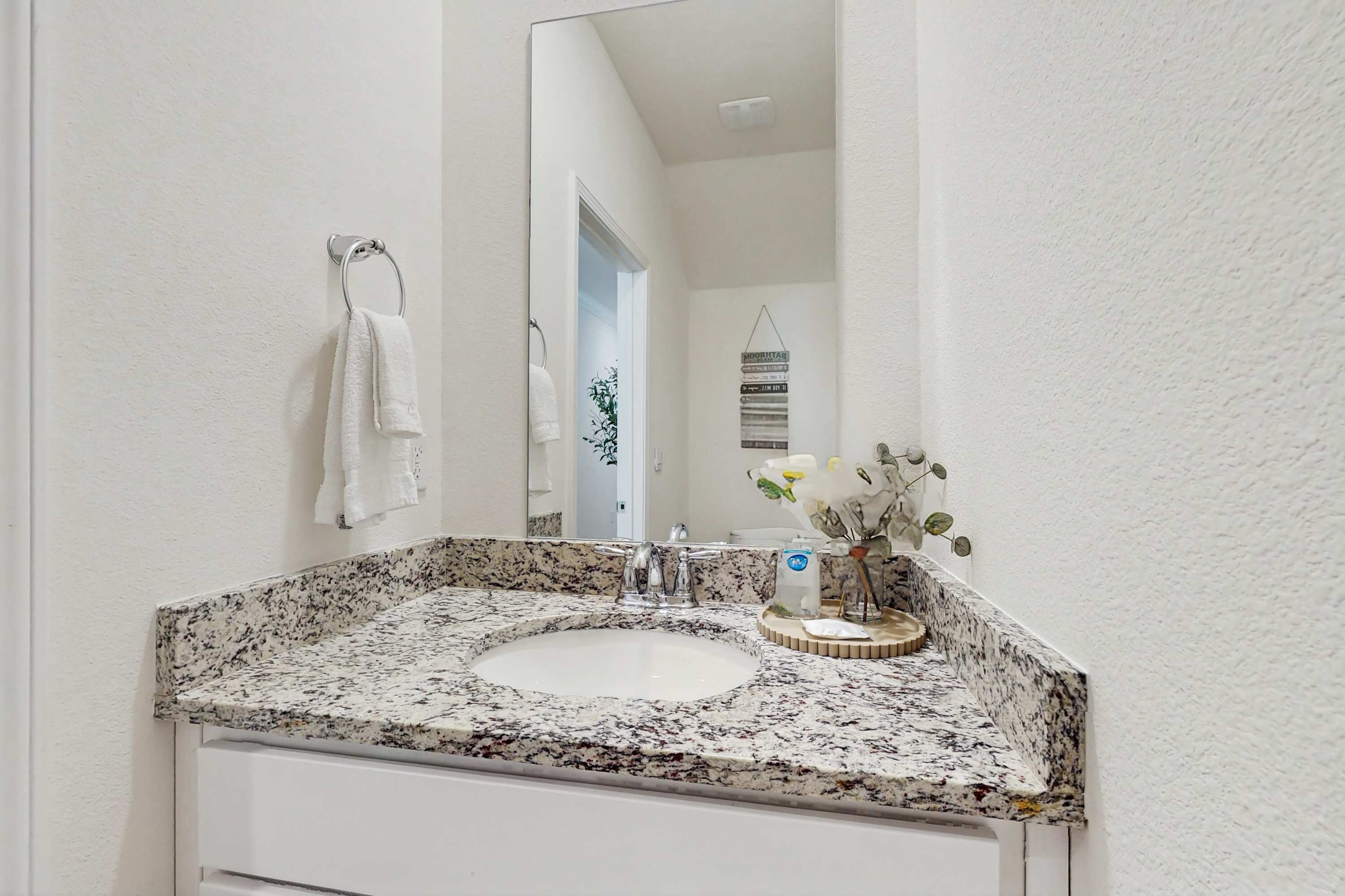 A bathroom with a granite countertop sink, a white towel hanging on a metal ring, and a mirror reflecting a door and a wall-mounted decoration.
