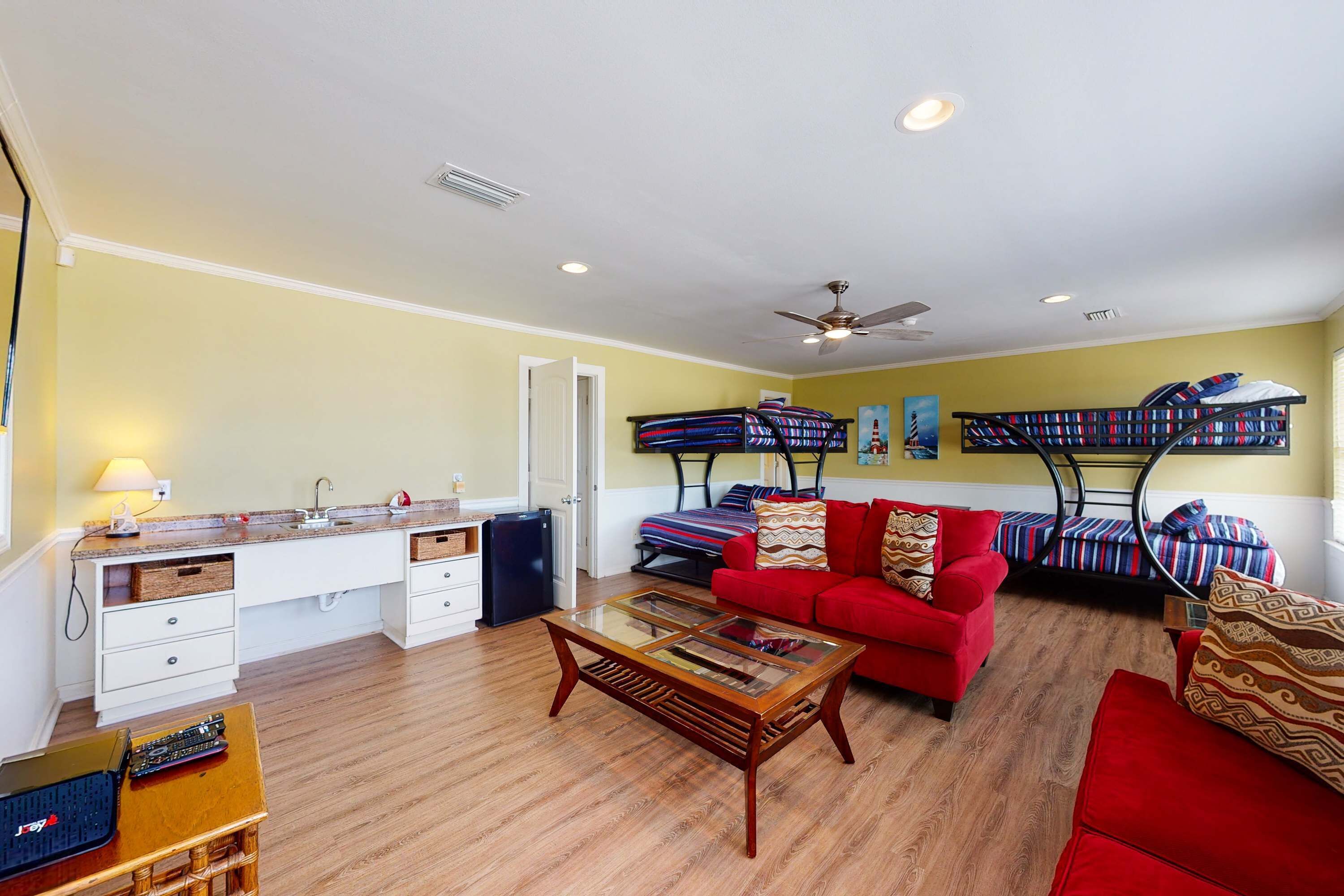 A compact living space with a kitchenette, a red sofa, a glass-top coffee table, and bunk beds with patterned bedding.
