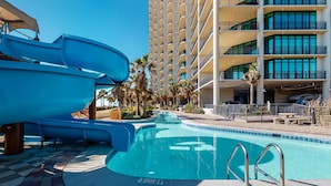 Una piscina con tobogán, palmeras y un edificio de varios pisos con balcones.