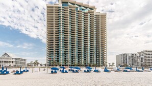 Un hotel alto con varios balcones, una playa con sillas azules para tomar el sol y un cielo despejado.