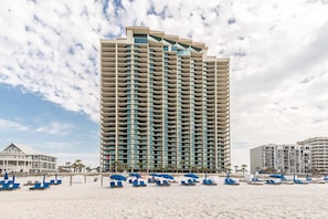 A tall hotel with multiple balconies, a beach with blue lounge chairs, and a clear sky. - Gulf front condo with direct beach access, resort amenities, private W&D, WiFi (Orange Beach)