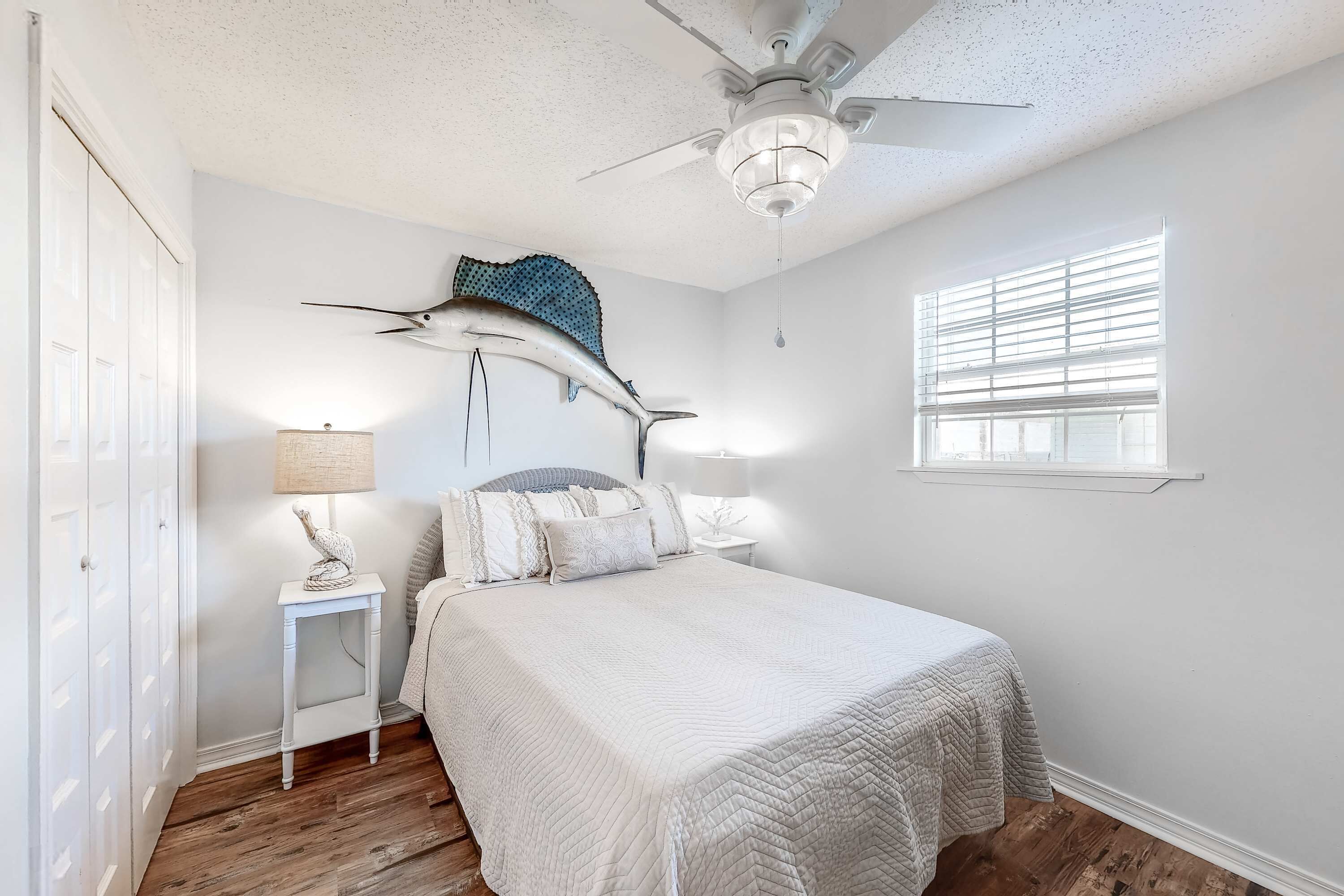 A bedroom with a bed, a ceiling fan, a window with blinds, and a wall-mounted lamp.