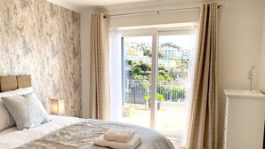Ein Schlafzimmer mit einem großen Bett, einem Nachttisch, einem Beistelltisch mit Lampe und Blick auf einen Balkon mit Geländer.