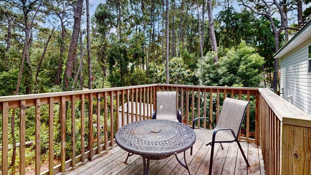 Une terrasse en bois avec une table ronde en métal et deux chaises, entourée d’une balustrade en bois, donnant sur une zone boisée.