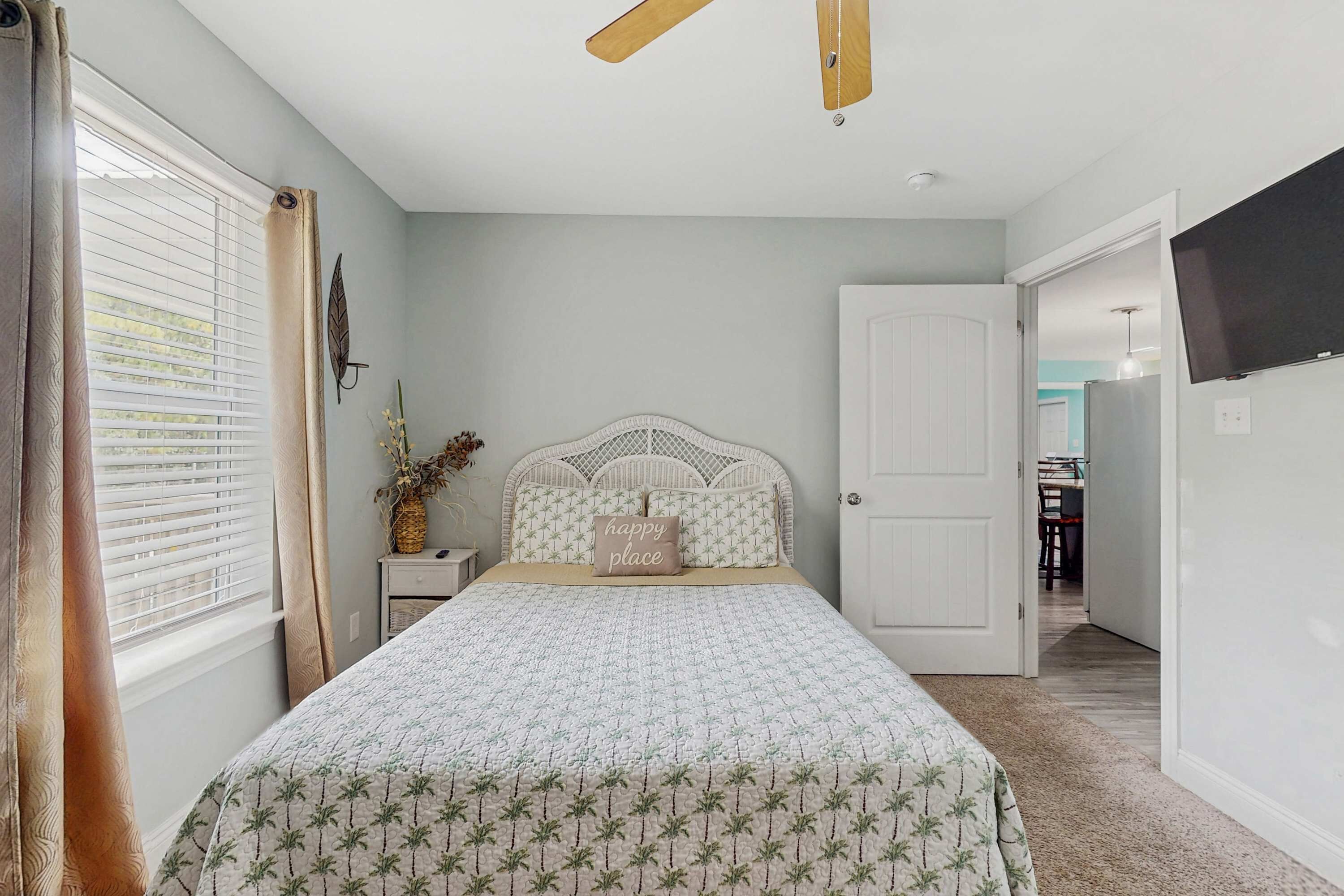A bedroom with a bed, a ceiling fan, a nightstand, a wall-mounted TV, and a door leading to another room.