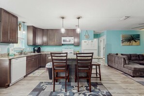 A modern kitchen with wooden cabinets, a microwave, and a dining area with two chairs. - Island Home With Spacious Deck, Lower Patio, Free WiFi, & Beach Access Nearby (Dauphin Island)