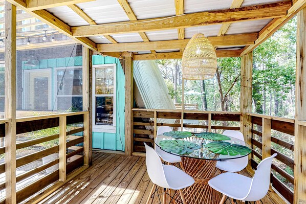 Une terrasse en bois avec une table en verre et des chaises blanches, entourée de rampes en bois et agrémentée d’un luminaire suspendu.