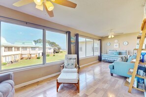 A spacious living area with large windows, a ceiling fan, a bed with striped bedding, and a blue sofa. - Family-friendly, beachfront getaway with a stainless kitchen & shared pool (Dauphin Island)
