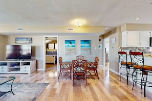 A modern living room with a flat-screen TV, wooden dining table, and chairs, a kitchen area with white cabinets, and artwork on the walls. - Family-friendly, beachfront getaway with a stainless kitchen & shared pool (Dauphin Island)