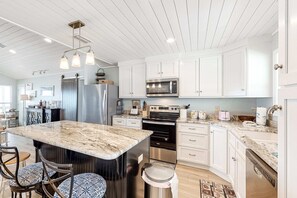 A kitchen with white cabinets, a marble countertop, stainless steel appliances, and a central island with bar stools. - Waterfront beach house on the sound with breathtaking sunset views & gulf access (Dauphin Island)