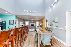 A spacious dining area with a long wooden table and chairs, a kitchen in the background, and a fireplace. - Bayfront home with wraparound deck & great water views - dog-friendly (Dauphin Island)