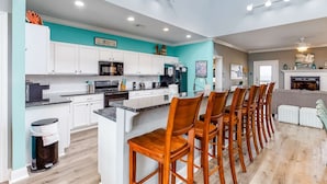 A modern kitchen with white cabinets, a central island with bar stools, and a dining area with a fireplace. - Bayfront home with wraparound deck & great water views - dog-friendly (Dauphin Island)