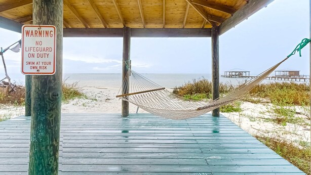 A wooden deck with a hammock, a warning sign, and a view of the beach. - Bayfront home with wraparound deck & great water views - dog-friendly (Dauphin Island)
