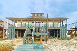 Una casa de playa de dos pisos, con balcón, terraza cubierta y escalera que lleva al nivel superior.