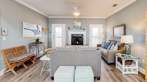 A living room with a grey sofa, wooden bench, and ottomans, a fireplace, and a ceiling fan. - Bayfront home with wraparound deck & great water views - dog-friendly (Dauphin Island)