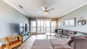 A living room with a sofa, a television, a ceiling fan, and a balcony view. - Bayfront home with wraparound deck & great water views - dog-friendly (Dauphin Island)
