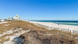 En strand med ett trästaket, en sandstig och en byggnad med flera våningar i bakgrunden.