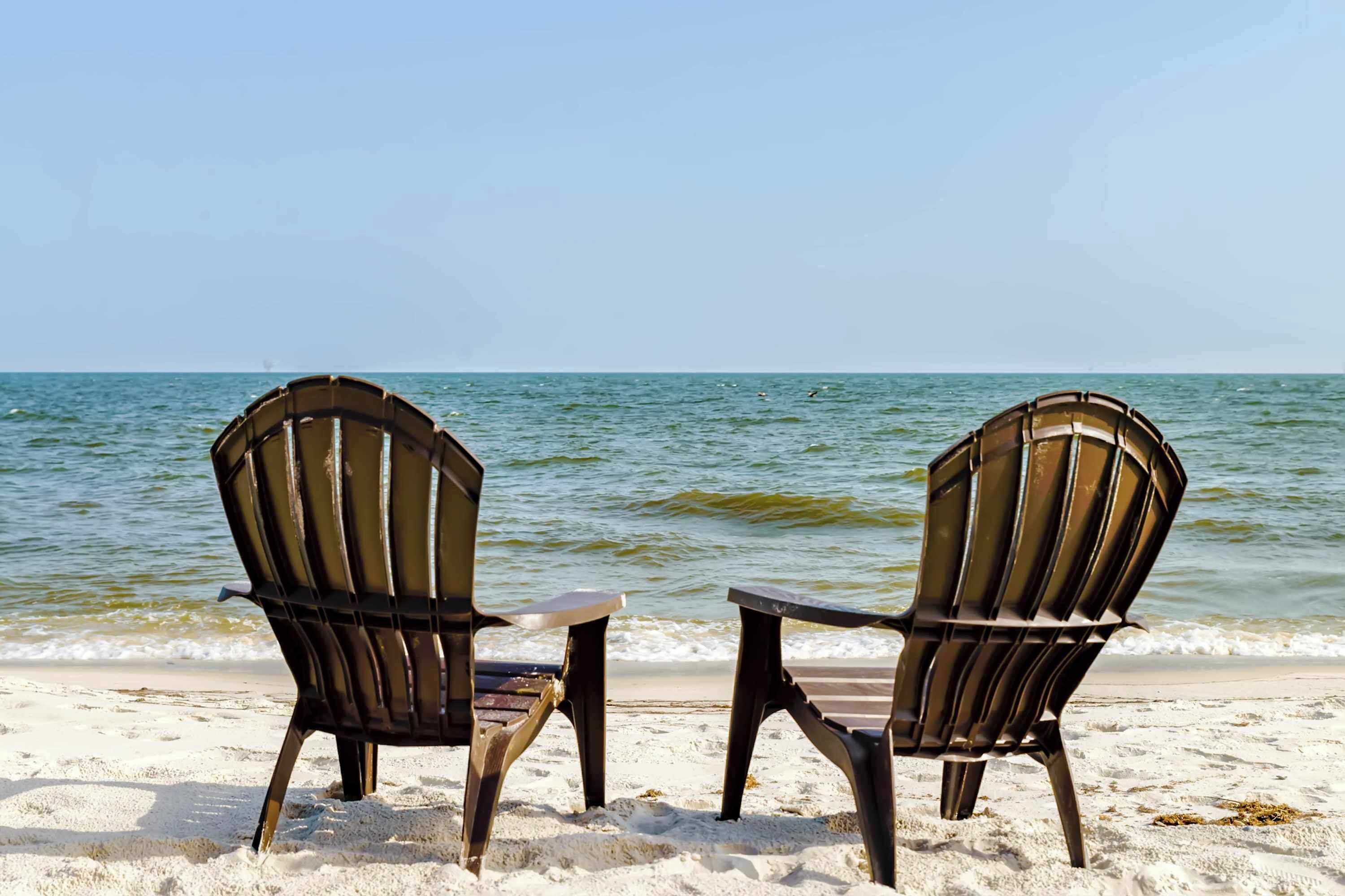 Two Adirondack chairs on a sandy beach with the ocean in the background.