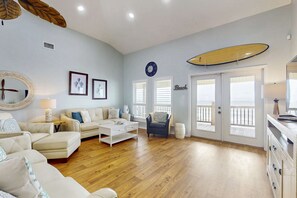 A spacious living room with a wooden floor, a white coffee table, a sofa, a chair, and a surfboard on the wall. - Beach Front house with breathtaking views, free WiFi, full kitchen (Dauphin Island)