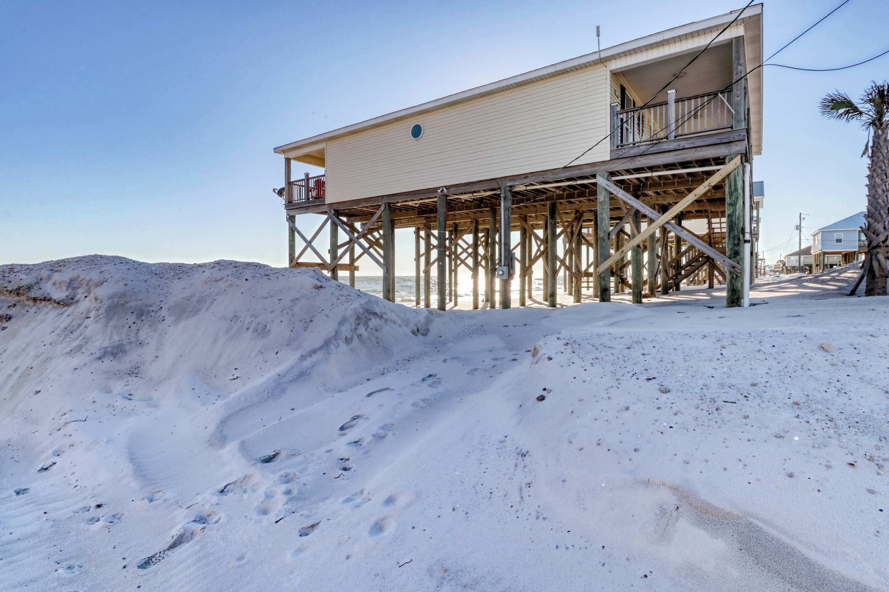 A beachfront house with a balcony, a sandy beach in front, and a palm tree to the right.