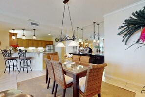 A dining area with wicker chairs, a glass table, and pendant lighting. - Bright beachfront condo with 2 Gulf-side pools, tennis courts & fitness room (Orange Beach)