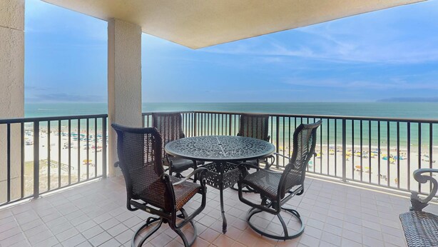 A balcony with a round table and four chairs, overlooking a beach with umbrellas. - Bright beachfront condo with 2 Gulf-side pools, tennis courts & fitness room (Orange Beach)