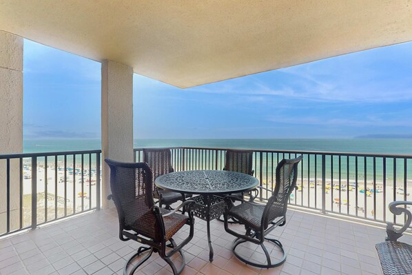 A balcony with a round table and four chairs, overlooking a beach with umbrellas. - Bright beachfront condo with 2 Gulf-side pools, tennis courts & fitness room (Orange Beach)