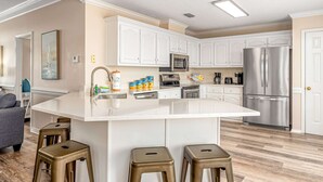A modern kitchen with white cabinetry, stainless steel appliances, and a central island with stools. - Newly remodeled bay view home with high-speed WiFi, central AC, and gas grill (Dauphin Island)