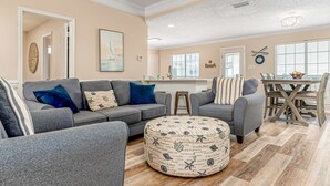 A living room with a gray sofa, ottoman, and a dining area with a wooden table and chairs. - Newly remodeled bay view home with high-speed WiFi, central AC, and gas grill (Dauphin Island)