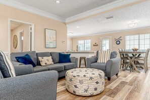 A living room with a gray sofa, ottoman, and a dining area with a wooden table and chairs. - Newly remodeled bay view home with high-speed WiFi, central AC, and gas grill (Dauphin Island)