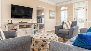 A living room with a flat-screen TV, grey sofas, a patterned ottoman, and a dining area with a white cabinet and a blue vase. - Newly remodeled bay view home with high-speed WiFi, central AC, and gas grill (Dauphin Island)