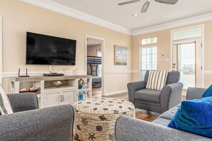 A living room with a flat-screen TV, grey sofas, a patterned ottoman, and a dining area with a white cabinet and a blue vase. - Newly remodeled bay view home with high-speed WiFi, central AC, and gas grill (Dauphin Island)