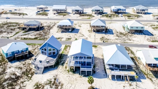 Une zone côtière comprenant plusieurs maisons de plage, dont certaines sont surélevées et d’autres ont des toits plats, le tout situé sur un terrain sablonneux, avec une route au premier plan.