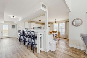 A spacious living area with a dining table, chairs, a kitchen in the background, and a large window with curtains. - Bay Front Home with - Gulf Views, WiFi, & Washer/Dryer - Two Dogs Welcome (Dauphin Island)