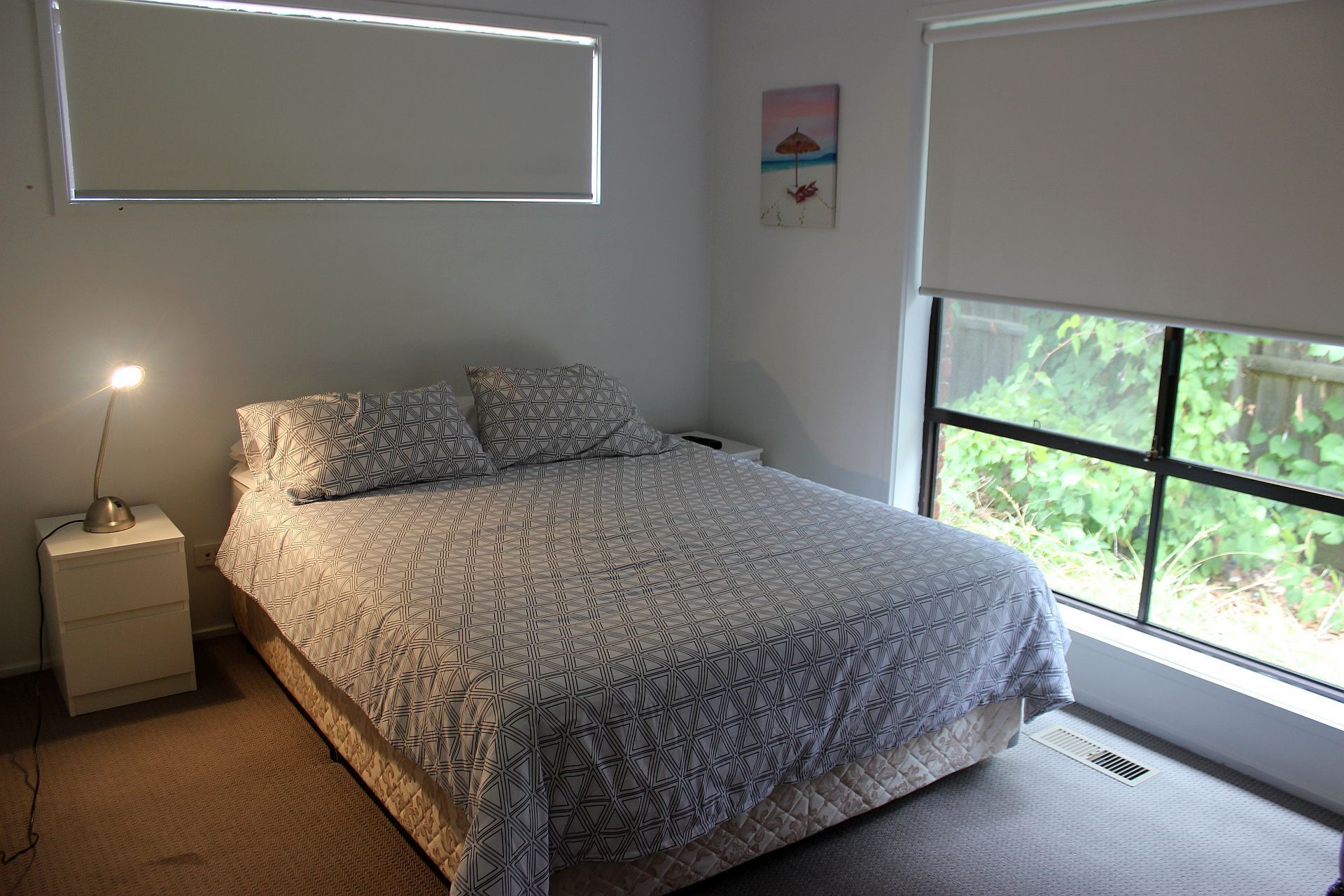 A bedroom with a bed, a nightstand, a lamp, a window with a view of greenery, and a painting on the wall.