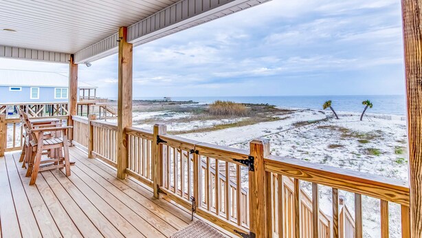 Una terraza de madera con barandilla, con vistas a una playa de arena y vegetación escasa.