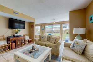 A living room with a sofa, armchairs, a coffee table, a TV, and a window with a view of buildings. - Condo with WiFi, boardwalk to beach, Gulf view, pool, & picnic area (Dauphin Island)