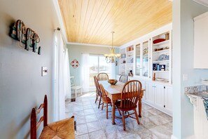 A dining area with a wooden table and chairs, a chandelier, and a kitchen with white cabinets and various dishes. - Charming bay-front home with WiFi, spacious wraparound porch, private deck (Dauphin Island)