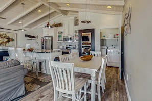 A spacious kitchen with white cabinetry, a dining area with a wooden table and chairs, and a high ceiling with exposed beams. - Charming beachfront home with Gulf views & wraparound deck - right on the sand (Dauphin Island)