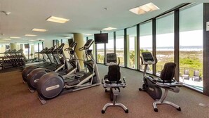 Une salle de sport équipée de tapis de course, de vélos d’appartement et d’appareils de musculation, de grandes fenêtres donnant sur la plage et d’un téléviseur à écran plat.