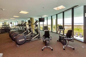 A gym with treadmills, stationary bikes, and weight machines, large windows with a view of the beach, and a flat-screen TV. - Beachfront condo with pool, private balcony, and washer and dryer (Dauphin Island)