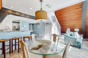 A modern kitchen with a glass dining table, wooden chairs, and a hanging light fixture. - Unique beachside A-frame with porches, fantastic views & ocean access (Dauphin Island)