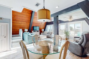 A spacious living room with a glass dining table, comfortable seating, a large wooden wall, and a ceiling fan. - Unique beachside A-frame with porches, fantastic views & ocean access (Dauphin Island)