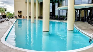 Un espace piscine avec une eau d’un bleu limpide, entouré de chaises longues et de tables, et de grandes fenêtres qui reflètent le ciel.