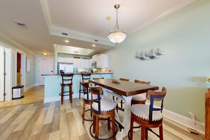A dining area with a wooden table and chairs, a kitchen in the background, and a light blue wall with sailboat decorations. - Beachfront condo with pool, private balcony, and laundry (Dauphin Island)