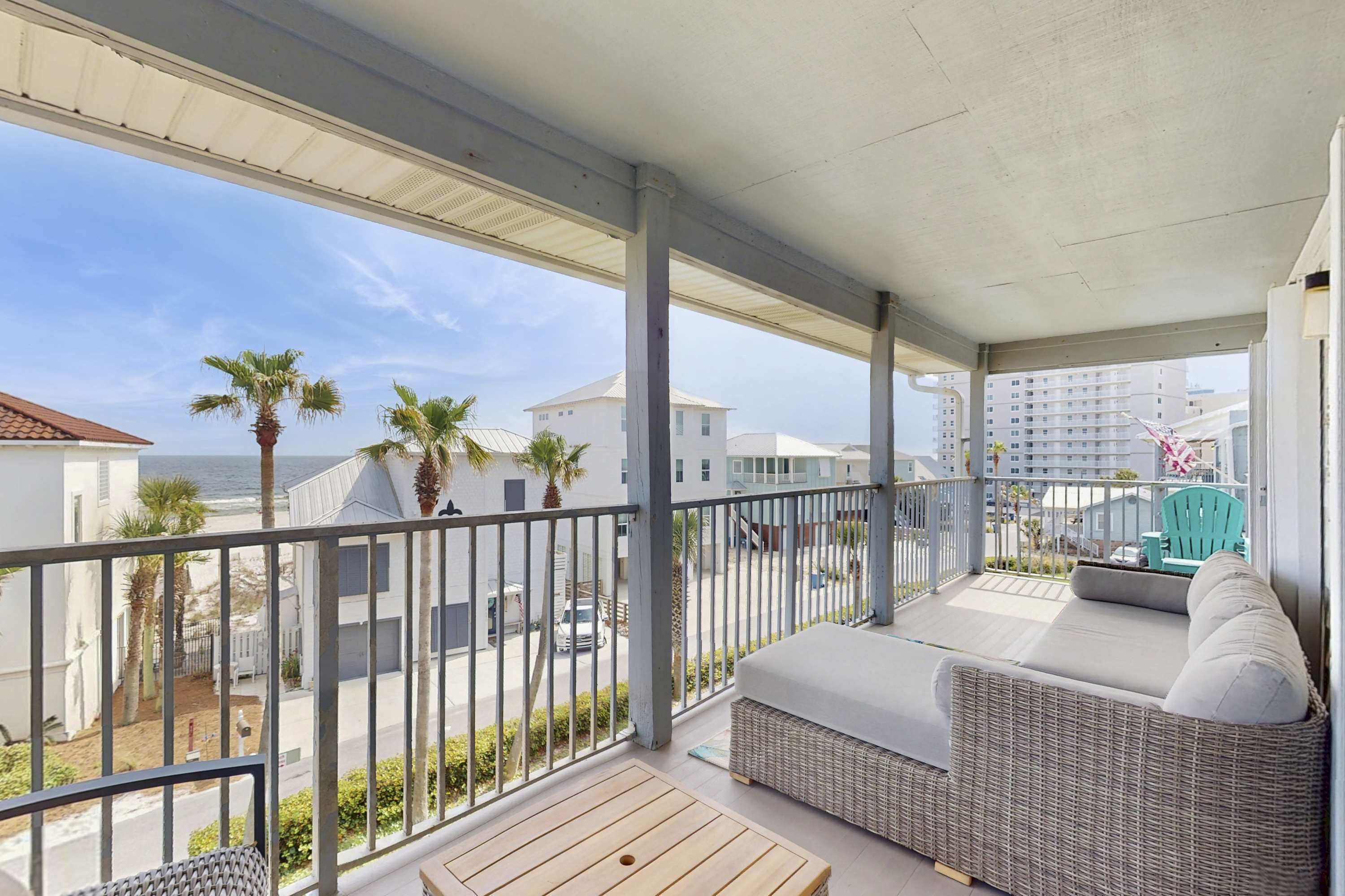 A balcony with a wicker sofa, a wooden table, and a view of the beach and buildings.