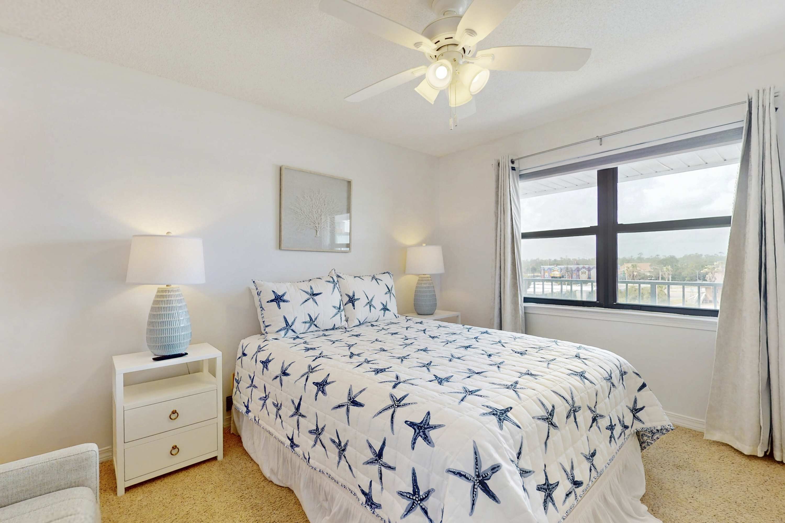 A bedroom with a bed, nightstands, a ceiling fan, a window with curtains, and a view of the outside.