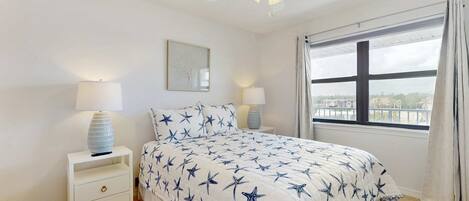 A bedroom with a bed, nightstands, a ceiling fan, a window with curtains, and a view of the outside.