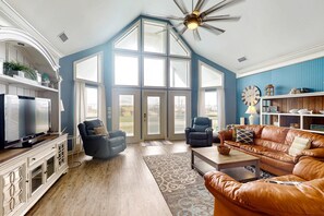 A spacious living room with a large window, a ceiling fan, a television, and a leather sofa. - Two-story, dog-friendly home with partial ocean views, decks, private washer,dryer (Dauphin Island)
