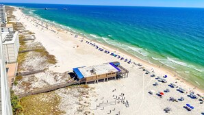 An aerial view of a beach with a lifeguard station, beach umbrellas, and a pier in the background. - Beachview condo with lazy river, marina, multiple pools and internet (Orange Beach)